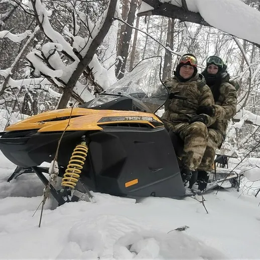 Два человека на снегоходе в зимнем лесу, окруженные снежными деревьями и холодной атмосферой