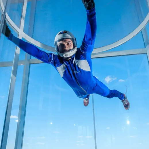 Человек в синем костюме выполняет свободное падение в аэродинамической трубе на фоне ясного неба
