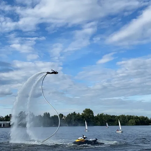 Человек на водном джет-паке взмывает в воздух на фоне облачного неба и озера, недалеко от зеленых деревьев