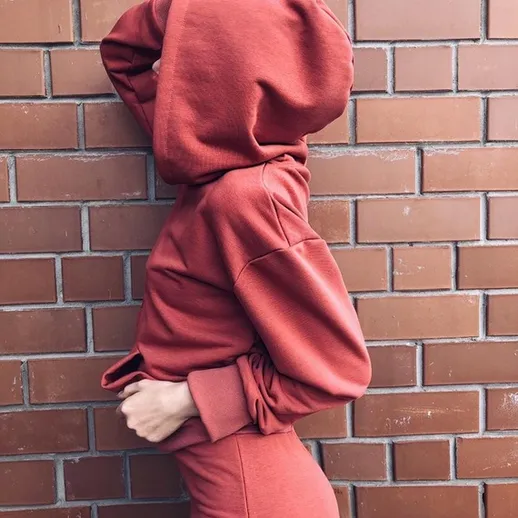 Молодая женщина в красном свитшоте стоит у кирпичной стены, позируя с загадочным выражением лица