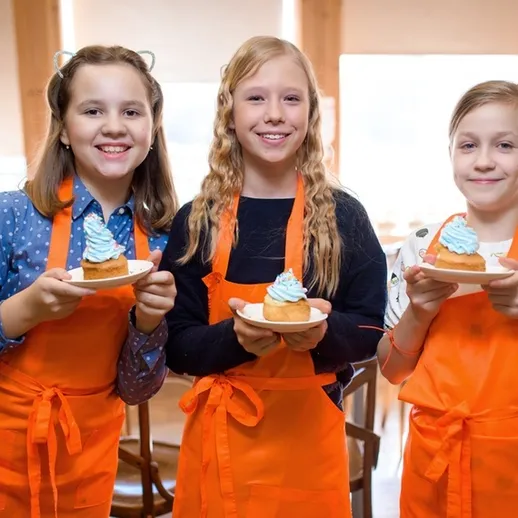 Три девочки в оранжевых фартуках с тортами на мастер-классе по кулинарии в уютном кафе