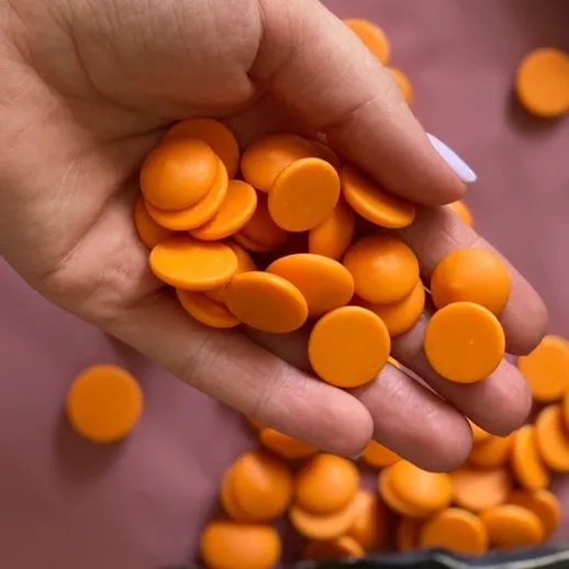 В руках держат много оранжевых капсул, рассыпанных на фоне ткани, вероятно, это конфеты или ингредиенты для выпечки