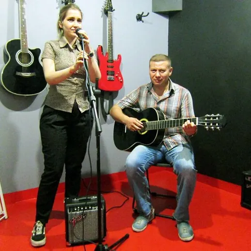 Женщина поет в репетиционной студии, а мужчина на гитаре аккомпанирует ей