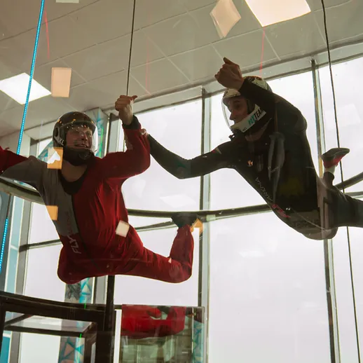 Два человека в аэродинамической трубе занимаются флайбордингом в помещении на фоне стеклянной стены