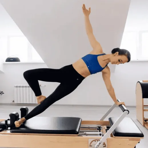 Женщина выполняет упражнение на реформере пилатеса в светлом зале с большими окнами и деревянной платформой