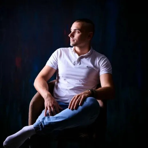 Молодой человек в белой футболке сидит на кресле с закрещенными ногами в полумраке