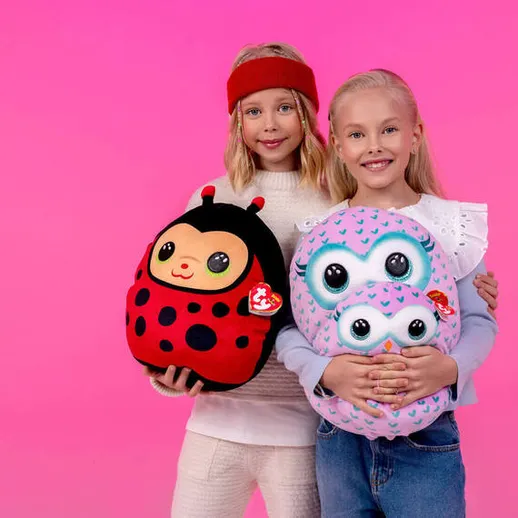 Две девочки улыбаются и держат мягкие игрушки на розовом фоне