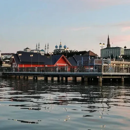 Спокойный вечер на набережной города у воды, с отражением зданий в реке под ясным небом