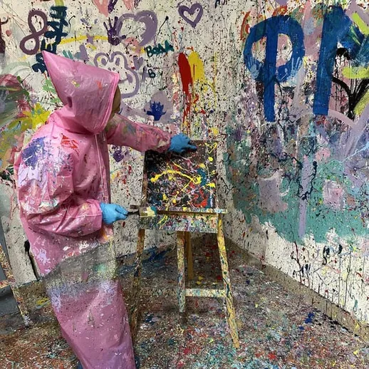 Художник в розовом комбинезоне пишет абстрактное произведение на мольберте в творческой мастерской с яркими стенами