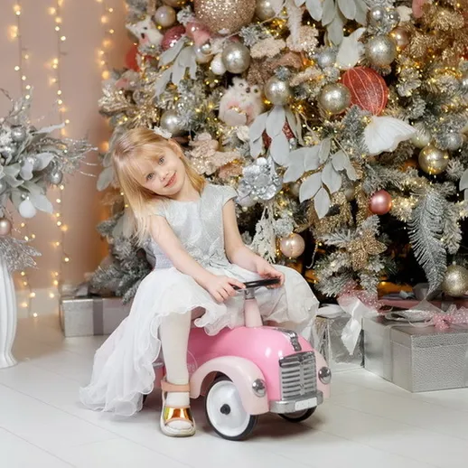 Девочка в белом платье весело сидит на розовой машинке перед рождественской елкой в уютном интерьере