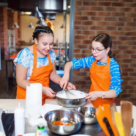 Две девочки в оранжевых фартуках готовят тесто на кухне в учебном классе кулинарии
