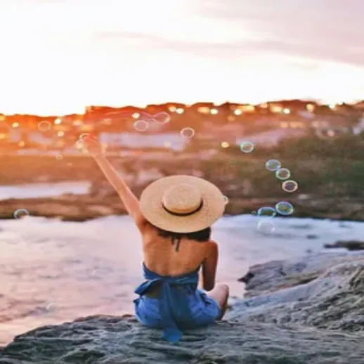 Девушка в шляпе сидит на скале у воды и запускает мыльные пузыри на фоне заката в прибрежном городке