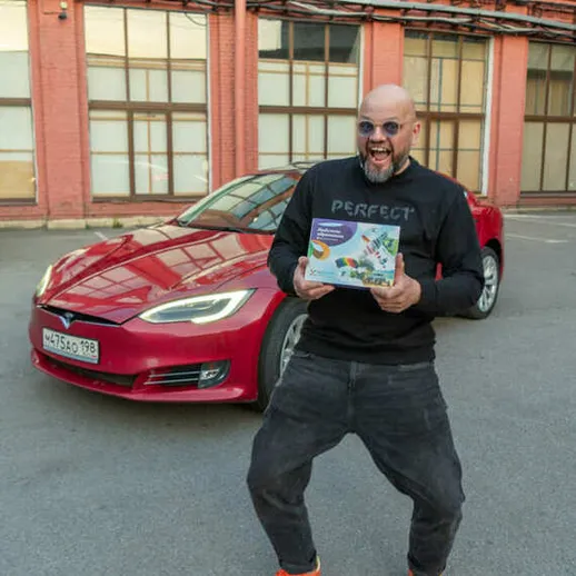Человек радостно позирует с коробкой подарка на фоне красного электромобиля во дворе здания
