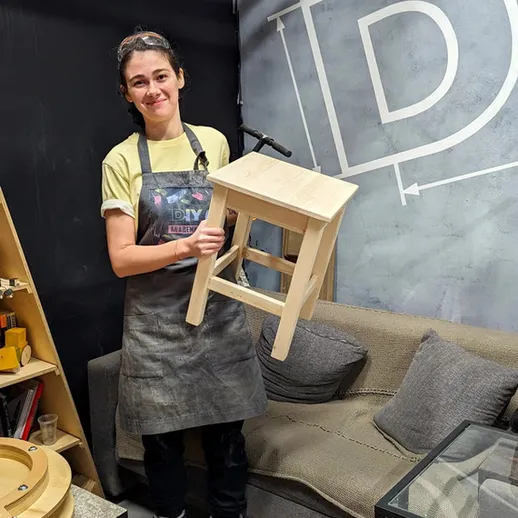 Женщина в рабочем фартуке держит деревянный стул в мастерской рядом с диваном и инструментами