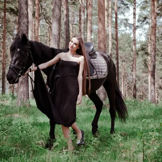 Молодая женщина в длинном платье ведет черную лошадь по лесу в солнечный день