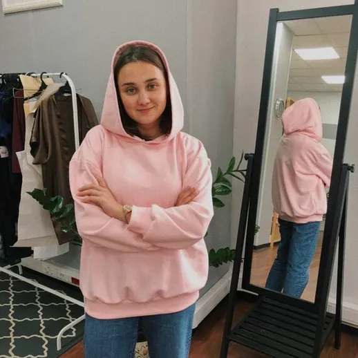 Молодая женщина в розовом худи стоит перед зеркалом в магазине одежды в светлом помещении