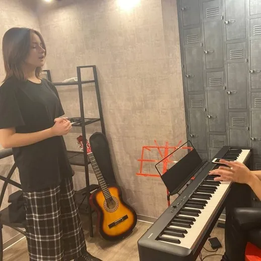 Девушка поет, а музыкант играет на клавишах в уютной комнате с гитарами и книжными полками