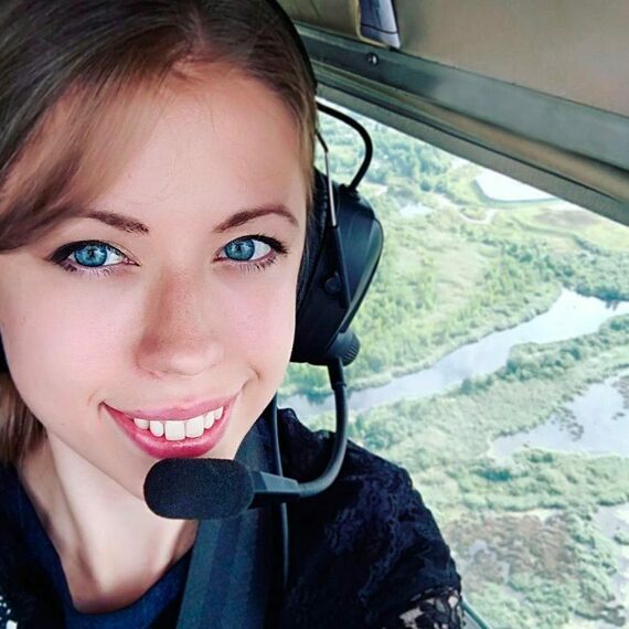 Девушка с блондинистым волосом в наушниках позирует в кабине вертолета над зелеными ландшафтами и реками. Фото 9