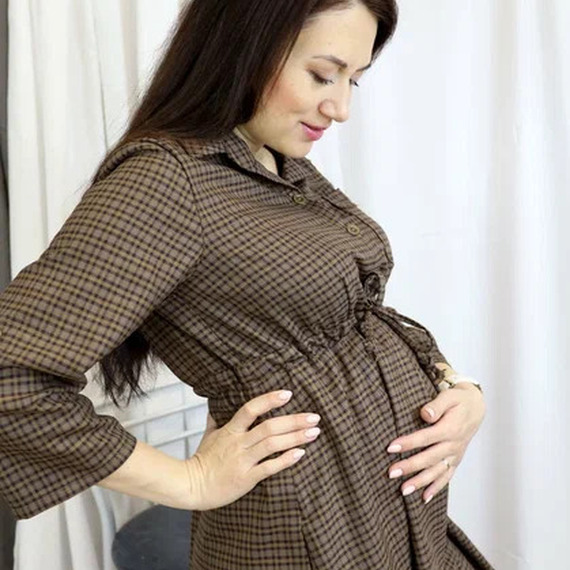 Беременная женщина в коричневом платье позирует на фоне белой ткани в уютной обстановке