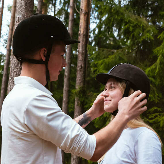 Молодой человек помогает девушке надеть шлем для верховой езды в лесу в солнечный день