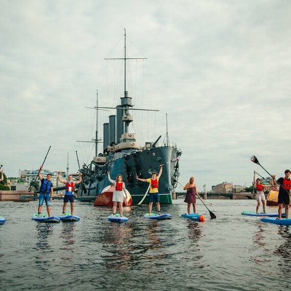 Группа людей на SUP-досках катается на воде в историческом центре, рядом с военным кораблем и современными зданиями