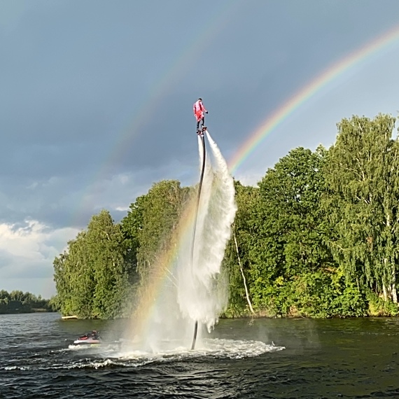 Человек на подъёмном устройстве в озере под радугой на фоне зелёных деревьев