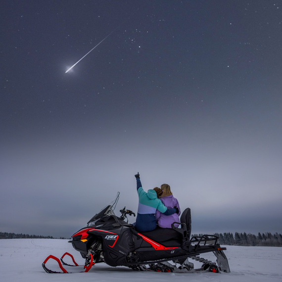Два человека на снегоходе смотрят на метеор в ночном небе над снежным полем