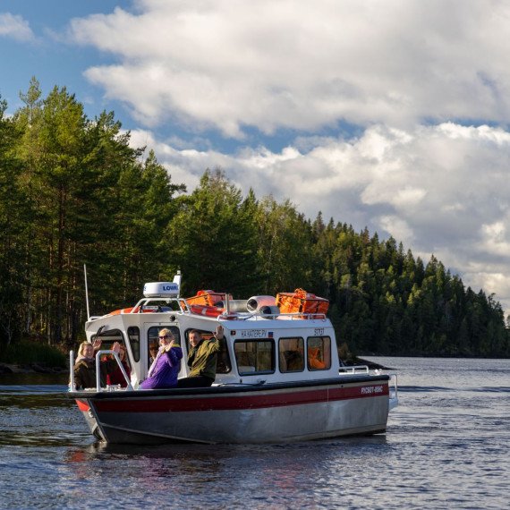 На реке плывёт катер с людьми среди зелёных деревьев и облачного неба