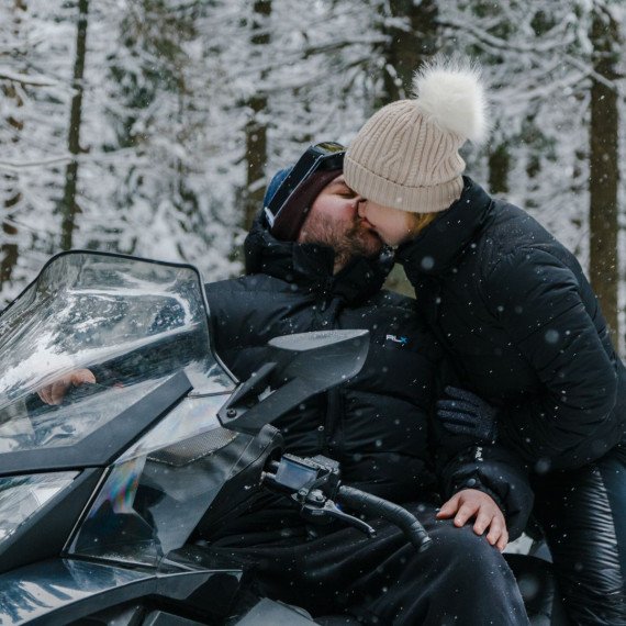 Пара на снегоходе целуется в зимнем лесу, вокруг снег и деревья, зима