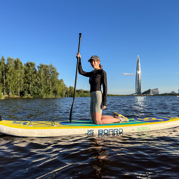 Девушка на SUP-доске катается по реке в ясный день, на фоне современного города и зелёного берега. Фото 12