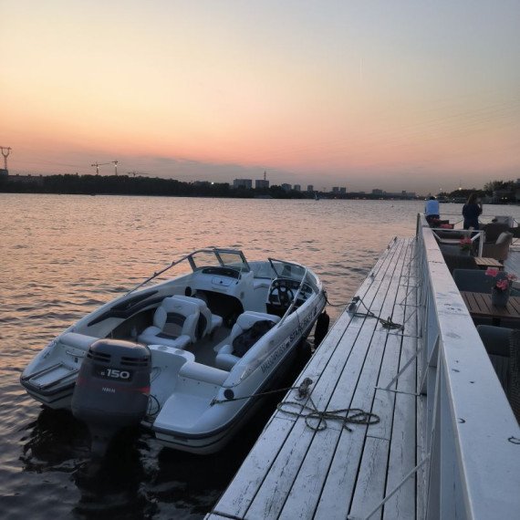 На реке стоят лодка и причал на фоне заката и городских зданий