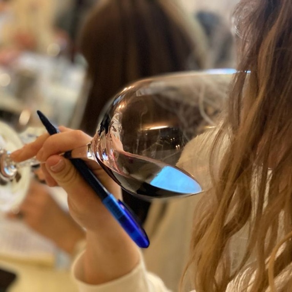 Участник винной дегустации в ресторане, внимательно оценивающий аромат вина, держа в одной руке бокал, а в другой - ручку