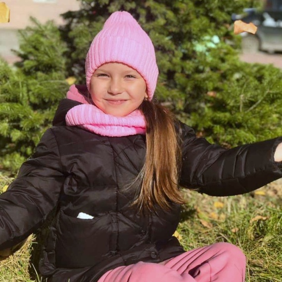 Девочка в розовой шапке и шарфе наслаждается осенним днем на улице, сидя на траве среди зелёных деревьев. Фото 9